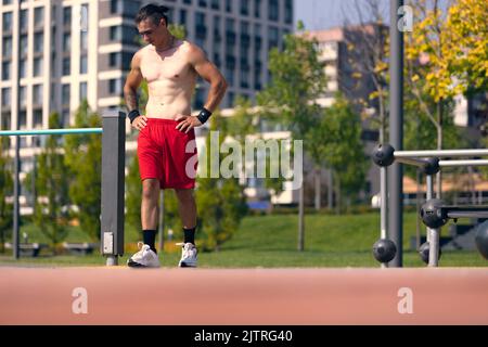 Beau jeune homme musclé en short rouge sur le terrain de sport du parc public en été matin. Sport, forme physique, concept de mode de vie sain Banque D'Images