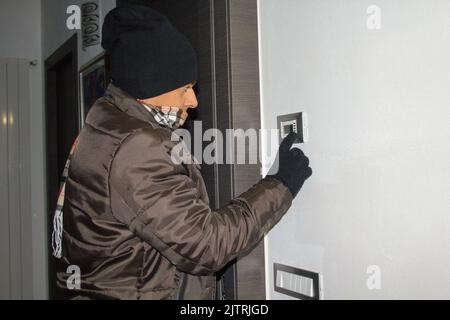 Homme en veste, écharpe, gants et chapeau de laine se sentant froid à la maison tout en retournant le thermostat de chauffage. Référence à l'hiver à venir Banque D'Images