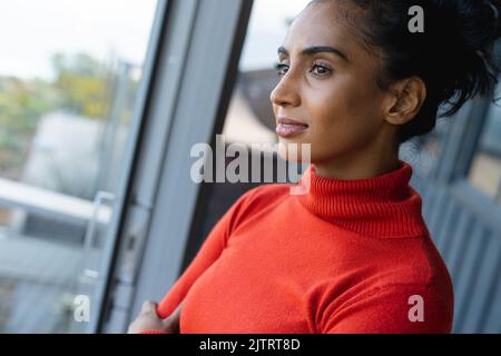 Gros plan d'une femme biraciale de taille moyenne, qui regarde par la fenêtre à la maison, espace pour le photocopie Banque D'Images