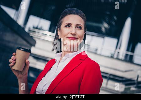 Photo de la charmante femme de secrétaire senior vêtue de blazer rouge appréciant pause-café en marchant à l'extérieur de la ville urbaine rue Banque D'Images