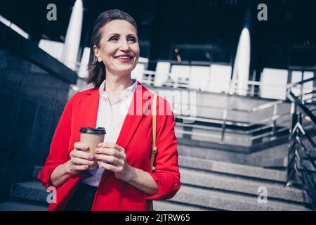 Photo d'une adorable femme de secrétaire sénior vêtue d'un blazer rouge en dégustant une boisson savoureuse en marchant à l'extérieur de la rue urbaine Banque D'Images
