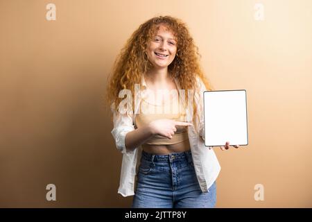 Modèle de femme souriante et souriante avec une coiffure en forme de curly dans des vêtements modernes pointant vers la tablette à écran blanc à l'aide d'un index. Banque D'Images