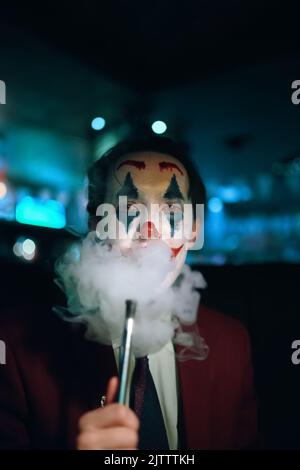 Guy en costume et cravate avec le Joker maquillage puffs fumée hors de sa bouche. Banque D'Images