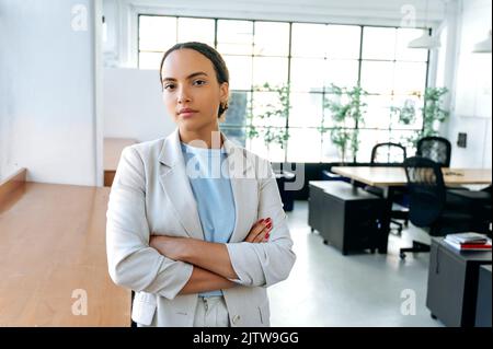 Portrait d'une jeune femme d'affaires de course mixte, forte d'une grande réussite, portant une tenue formelle, debout dans un bureau moderne avec des bras croisés, regardant l'appareil photo Banque D'Images