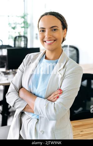 Photo verticale d'une jeune femme d'affaires élégante et prospère, avec un sourire blanc parfait, en tenue formelle, dans un bureau moderne avec ses bras croisés, regardant l'appareil photo, souriant Banque D'Images