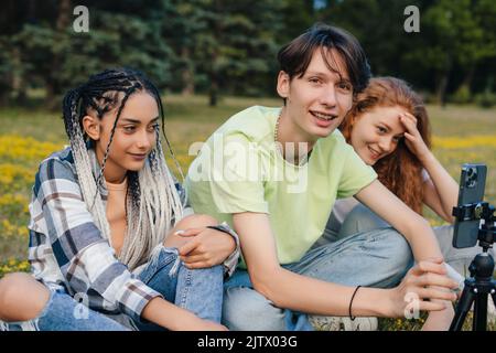 Un groupe de trois personnes utilisant un trépied et un téléphone pour prendre une photo de groupe pendant qu'ils sont assis sur l'herbe dans le parc d'été. Concept de communauté avec Banque D'Images