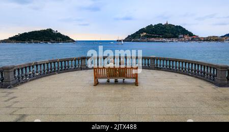 Couple non identifié sur un banc de promenade Paseo de la Concha avec vue sur la baie de la Concha et l'île de Santa Klara ou Santa Clara à San Sebastian, Espagne Banque D'Images