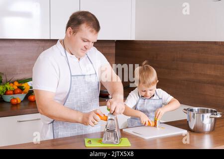 Le père cuisine avec son fils, frotte des carottes sur le râleur à la maison dans la cuisine, s'amuser ensemble, le père enseigne à son fils comment cuisiner, l'enfant aide heureusement Banque D'Images