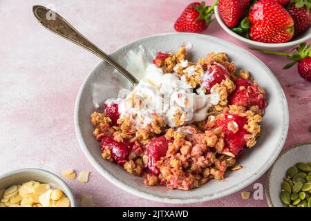 Délicieux crumble aux fraises avec de la crème glacée, des amandes et des graines de citrouille dans une assiette avec une cuillère sur fond rose. Gros plan. Petit déjeuner savoureux Banque D'Images
