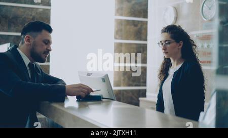 Homme d'affaires invité à la réception de l'hôtel, facture payante avec smartphone via la technologie de paiement sans contact et conversation avec le réceptionniste debout à la réception. Business, Travel et People concept Banque D'Images
