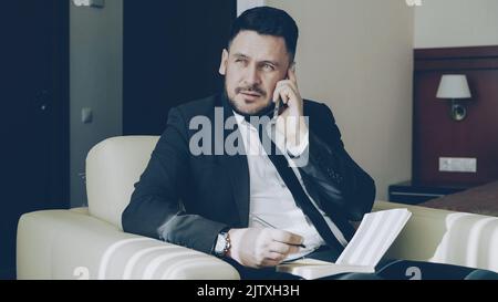 homme d'affaires confiant parlant un téléphone portable et écrivant des notes dans le bloc-notes tout en étant assis sur un fauteuil dans la chambre d'hôtel. Concept de voyage, d'affaires et de personnes Banque D'Images