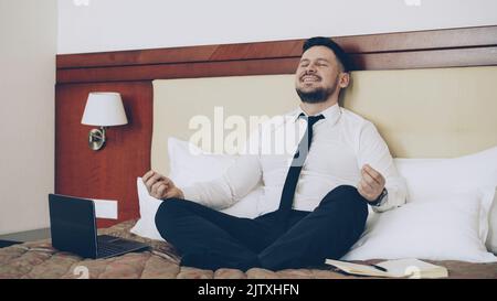 Un homme d'affaires heureux a mis un ordinateur portable et a pris la position yoga lotus assis sur le lit dans la chambre d'hôtel. Concept de voyage, d'affaires et de personnes Banque D'Images