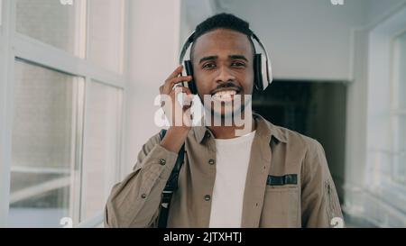 Portrait d'un jeune beau étudiant d'origine afro-américaine debout dans un large couloir blanc spacieux de l'université avec de grands écouteurs sur son cou, regardant l'appareil photo et souriant. Banque D'Images