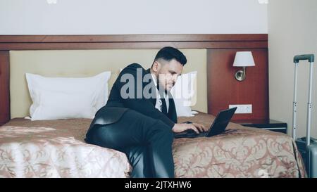 Photo panoramique d'un jeune homme d'affaires concentré en costume travaillant sur un ordinateur portable tout en étant assis sur un lit dans la chambre d'hôtel. Business, Travel et People concept Banque D'Images