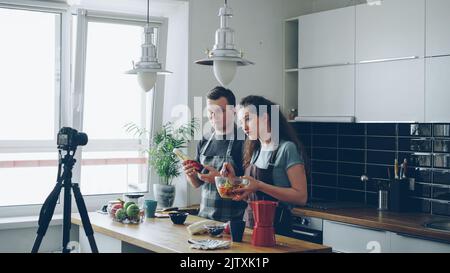 Un couple souriant et séduisant enregistre des vidéos sur la préparation de salade haelthy sur un appareil photo numérique dans la cuisine à la maison. Vlogging et concept de médias sociaux Banque D'Images