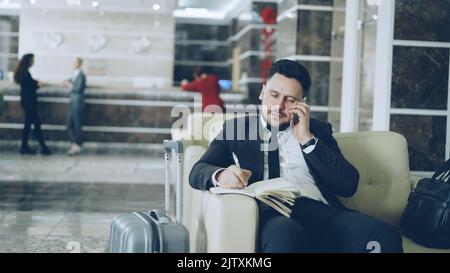 Prise de vue panoramique d'un homme d'affaires confiant parlant un téléphone portable et écrivant des notes dans un bloc-notes tout en étant assis sur un fauteuil dans un hôtel de luxe avec des bagages près de lui. Concept de voyage, d'affaires et de personnes Banque D'Images