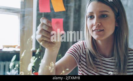 Gros plan d'une jolie jeune femme aux cheveux blonds collés des notes de couleur sur une planche en verre dans un bureau moderne. Elle regarde les journaux et sourit. Concept de travail créatif. Banque D'Images