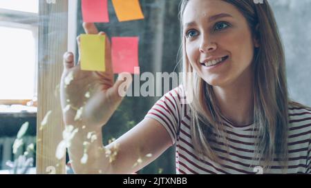 Gros plan d'une jolie jeune femme aux cheveux blonds collés des notes de couleur sur une planche en verre dans un bureau moderne. Elle regarde les journaux et sourit. Concept de travail créatif. Banque D'Images