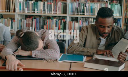 les étudiants diligent se préparant aux examens faisant leurs devoirs et un gars fatigué dormant sur une table dans la bibliothèque de l'université à l'intérieur Banque D'Images