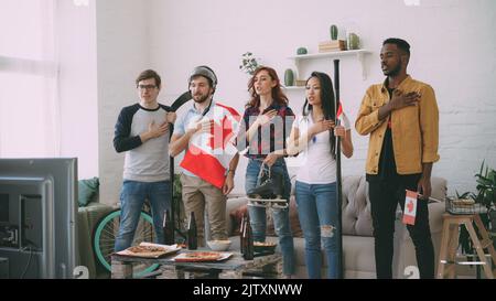 Groupe multiethnique d'amis amateurs de sport écoutant et chantant l'hymne national canadien avant de regarder le championnat sportif à la télévision ensemble à la maison à l'intérieur Banque D'Images