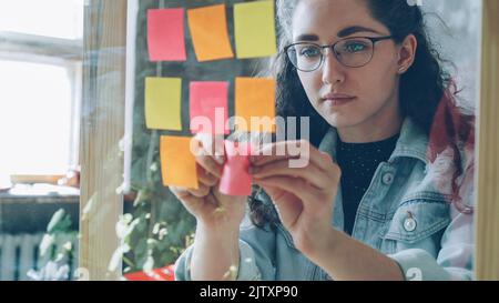 Gros plan d'une jeune femme portant des lunettes qui collent des notes de couleur sur une planche en verre dans un bureau moderne de style loft. Elle regarde des notes lumineuses et sourit. Banque D'Images