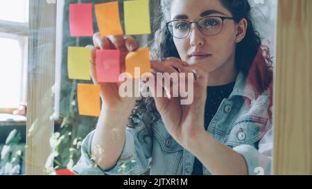 Gros plan d'une jeune femme portant des lunettes qui collent des notes de couleur sur une planche en verre dans un bureau moderne de style loft. Elle regarde des notes lumineuses et sourit. Banque D'Images