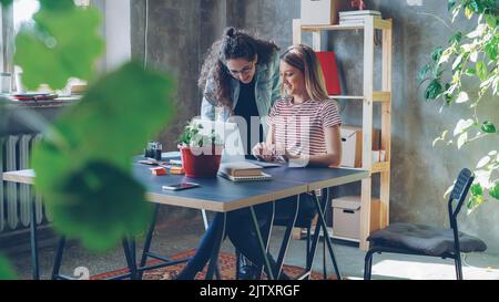 Les jeunes propriétaires de petites entreprises travaillent avec un ordinateur portable dans un bureau moderne de style loft. Blonde est assis et dactylographiée, brunette est debout près du bureau et suggère des idées regardant l'écran. Banque D'Images