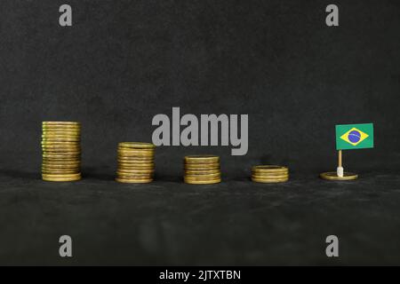 Concept de récession économique, crise financière et dépréciation du peso au Brésil. Drapeau brésilien dans la pile décroissante de pièces sur fond noir foncé. Banque D'Images