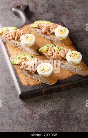 Toast de seigle avec thon en conserve, œuf dur, avocat mûr et graines de sésame sur une planche de bois sur la table. Verticale Banque D'Images