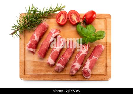 Rouleaux de steak de cou de porc cru (coppa) sur planche à découper en bois avec romarin, tomates cerises hachées et feuilles de basilic isolées sur blanc, vue de dessus, coupe-cheveux Banque D'Images