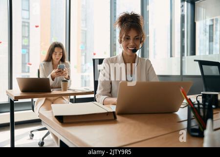 Une femme souriante avec un casque travaille à l'ordinateur portable, l'employé est assis derrière lui Banque D'Images