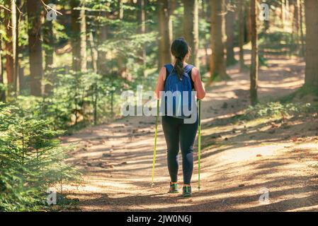 Femme marchant le long d'un chemin de forêt, tandis que les rayons du soleil se brisent à travers les arbres. Porte un sac à dos et des bâtons de randonnée Banque D'Images