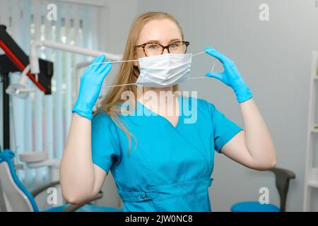 Bonne femme blanche dentiste en blouse de laboratoire bleue et masque protecteur posant avec un joli sourire en regardant la caméra dans son cabinet dentaire Banque D'Images