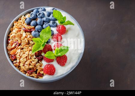 Granola avec yaourt et baies sur fond brun foncé. Petit déjeuner sain avec yaourt, granola cuite au four, myrtilles et framboises. Vue de dessus. Copier Banque D'Images