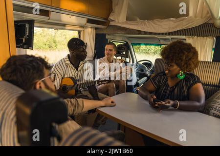 Groupe interracial actif de quatre bons amis sur une route d'été, assis à l'intérieur de leur confortable motorhome moderne, jouer de la guitare, bavarder et rire. Intérieur du RVS. Photo de haute qualité Banque D'Images