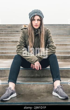 Belle jeune femme dans des vêtements décontractés tendance est assis sur des marches près de l'extérieur Banque D'Images