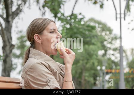 Une femme d'âge moyen est assise sur un banc dans le parc et mange de la crème glacée. Banque D'Images