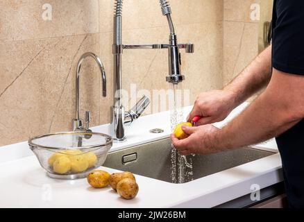 Un homme pele des pommes de terre sur la peau dans la cuisine et les met dans un panier Banque D'Images