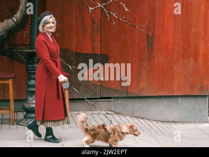 Femme âgée marchant avec un animal de compagnie charmant en pelage rouge. Vieille dame âgée à la retraite sur la rue de la ville avec des fleurs.personne aînée élégante retraité Banque D'Images
