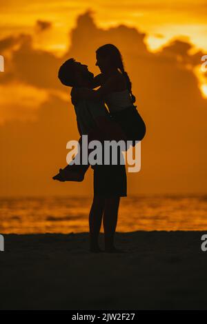 Vue latérale silhouettes de copain amoureux heureux levant petite amie souriante en se tenant sur la plage de sable près de l'océan onduleux sous le ciel lumineux de coucher de soleil à Cl Banque D'Images