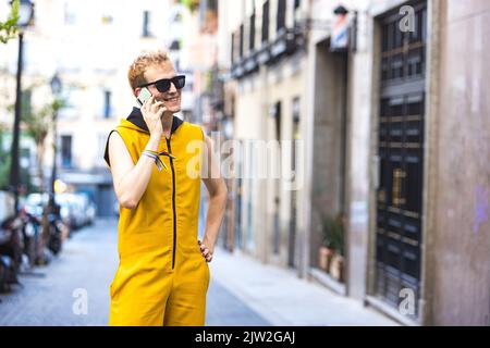 Joyeux homme transgenre jeune dans la combinaison jaune et les lunettes de soleil regardant loin en marchant parlant sur le téléphone portable dans la rue de ville Banque D'Images