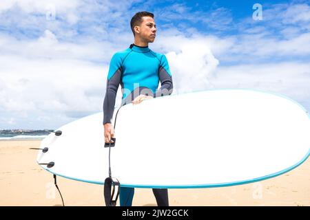 Jeune homme hispanique réfléchi surfeur avec cheveux foncés et en combinaison avec planche de surf blanche sur la plage de sable Banque D'Images
