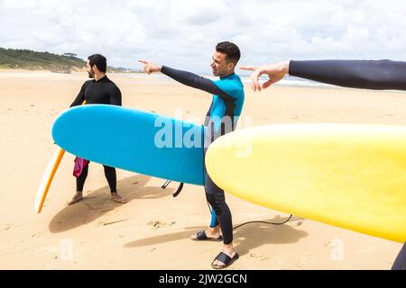 Corps complet de jeunes surfeurs masculins de forme ethnique avec des cheveux sombres dans des combinaisons debout sur la plage de sable près de l'océan avec des planches de surf colorées et pointant vers l'extérieur Banque D'Images