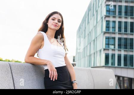 Magnifique jeune femme aux longs cheveux bruns dans une tenue élégante en souriant regardant l'appareil photo contre les gratte-ciel modernes de la ville Banque D'Images
