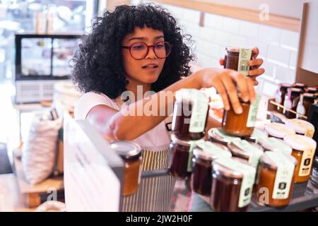 D'en haut jeune Afro-américaine vendeur de verres arranger des pots en verre avec diverses sauces à confiture sur les étagères pendant le travail dans les épiceries écologiques Banque D'Images