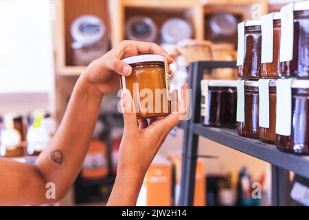 Une vendor féminin non reconnaissable en culture qui organise des bocaux en verre avec différentes sauces à confiture sur les étagères pendant le travail dans une épicerie écologique Banque D'Images