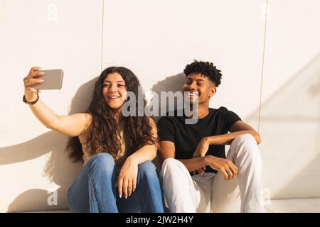 Un jeune couple souriant prend le selfie à travers un smartphone tout en étant assis près du mur Banque D'Images