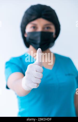Contenu jeune femme médique en bleu frotte gants de protection masque montrant les pouces vers le haut mouvement et regardant l'appareil photo contre fond blanc Banque D'Images