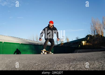 Hippster mâle barbu qui fait du skateboard le long de la piste de pompe tout en passant du temps dans le parc de skate par temps ensoleillé Banque D'Images