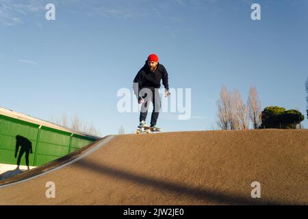 Hippster mâle barbu qui fait du skateboard le long de la piste de pompe tout en passant du temps dans le parc de skate par temps ensoleillé Banque D'Images